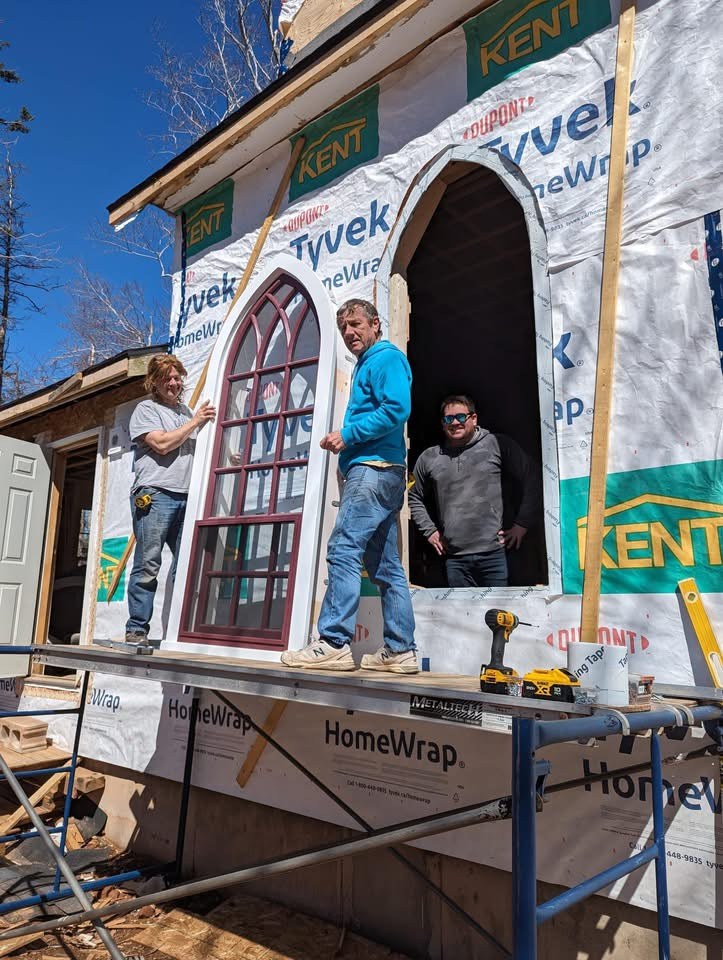 An arched window unit being set into a house under construction, scaffolding up and house wrap still on the walls