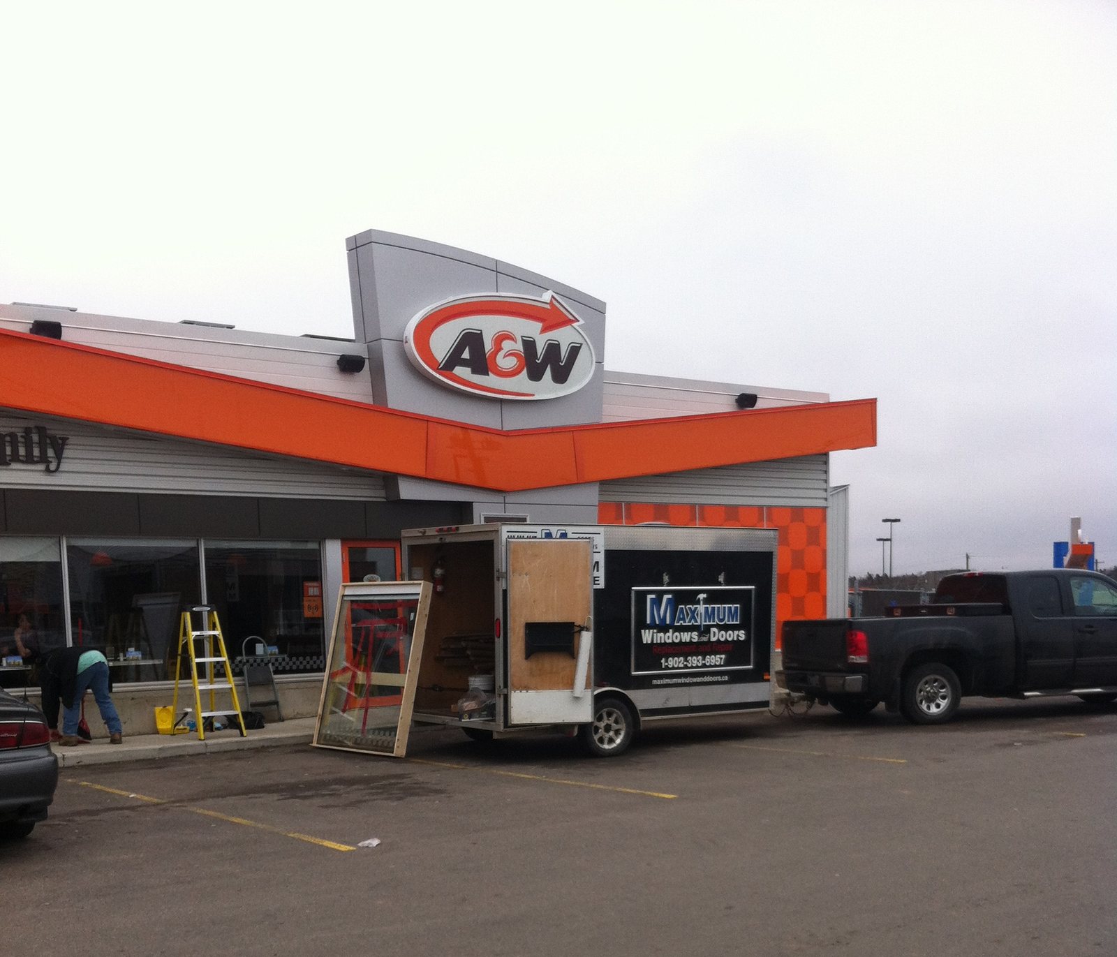 The Maximum work trailer parked at a restaurant storefront, a window unit leaning against it and a stepladder up while the glass is worked on